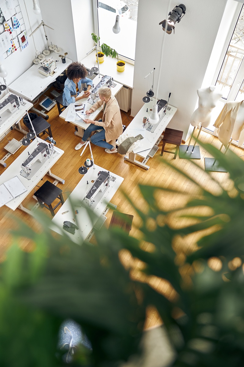 Short haired woman designer and African-American seamstress look at drawing in sketrchbook shooting video for blog at sewing machine in light fashion studio